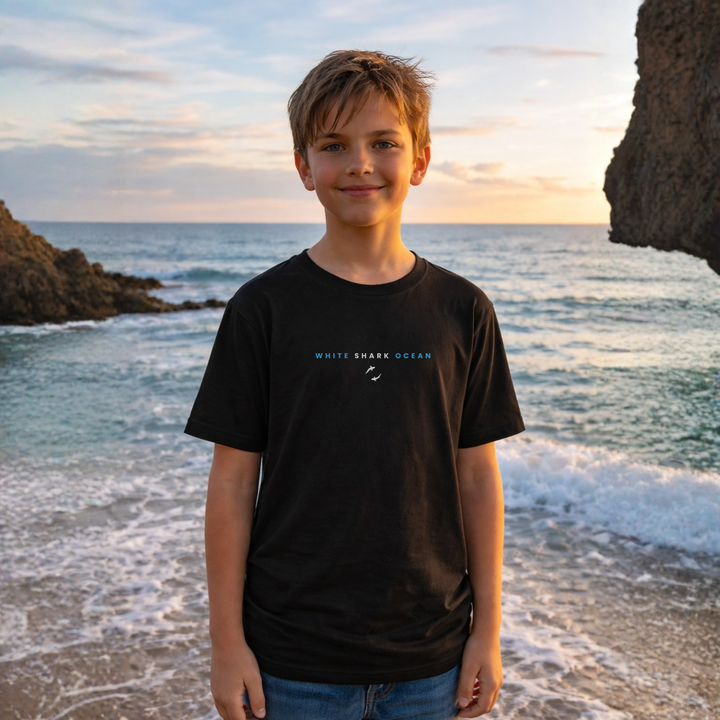 Young boy standing on a beach with ocean waves and rocks in the background, wearing a black t-shirt with white shark ocean logo.