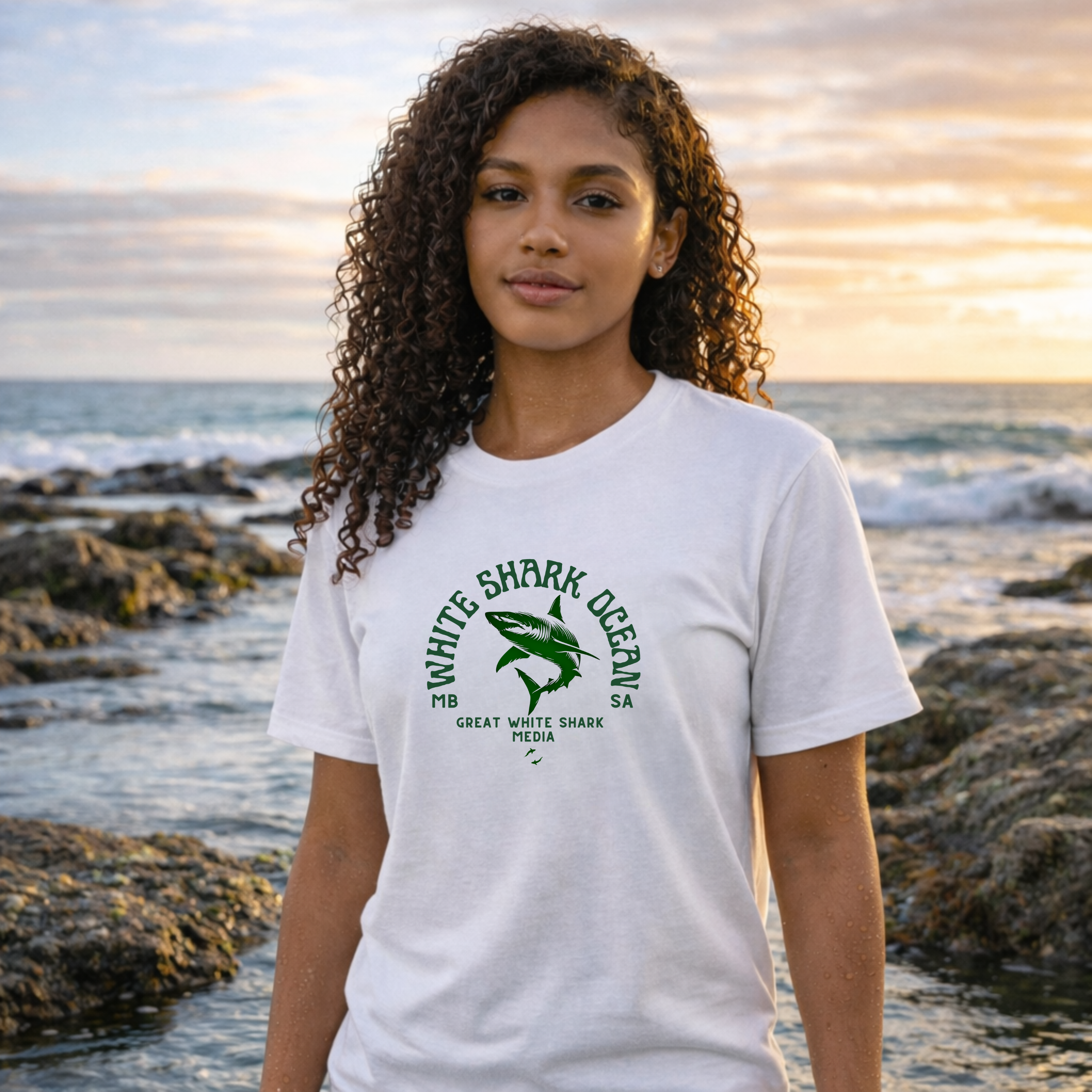 Person wearing a white t-shirt with a green graphic and text, standing on a rocky beach with ocean and sunset in the background.