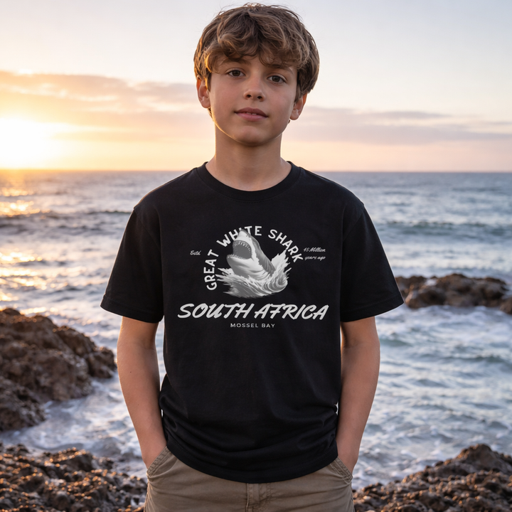 Young boy wearing a black t-shirt with a Great White Shark design and text, standing on a rocky beach at sunset. white shark ocean great white shark south africa
